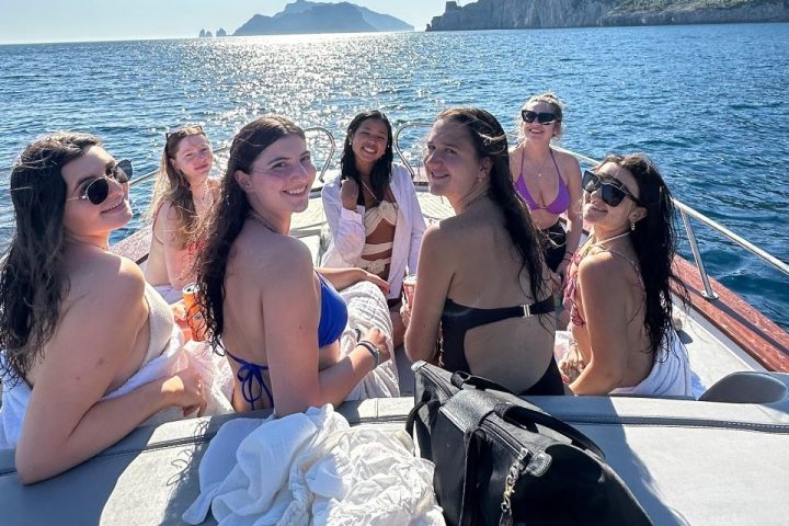 Six women on a boat in swimsuits, with sunny sky and coastal cliffs in the background.