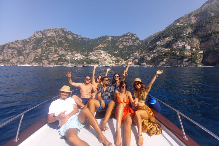 Group of six people on a boat, smiling and raising arms, with mountains and coastal town in the background.