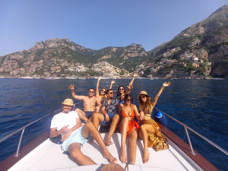 Group of six people on a boat, smiling and raising arms, with mountains and coastal town in the background.
