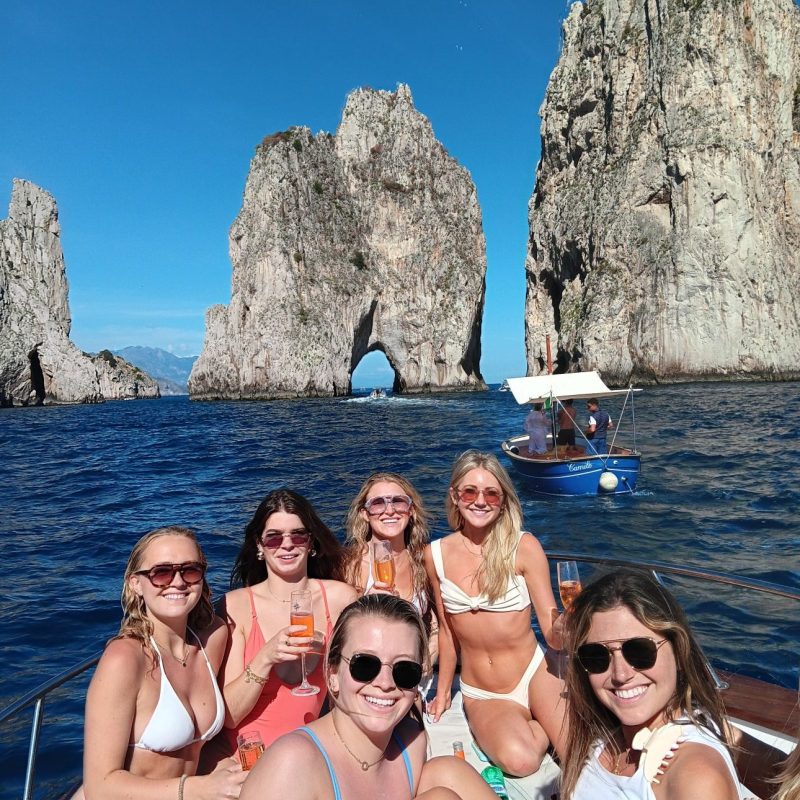 Six women on a boat in swimsuits, holding drinks, with rocky cliffs in the background.