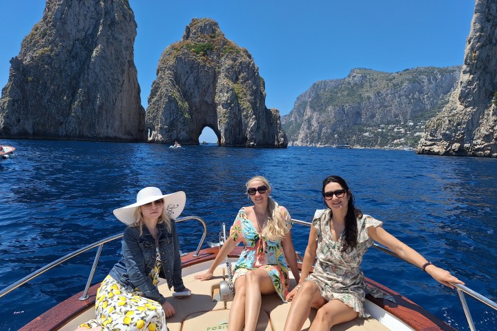 Three women sitting on a boat with rocky cliffs and blue sea in the background under a clear sky.