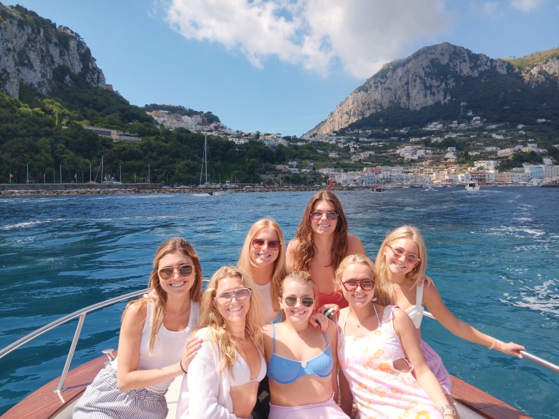 Seven women on a boat smiling, with a mountainous coastline in the background.