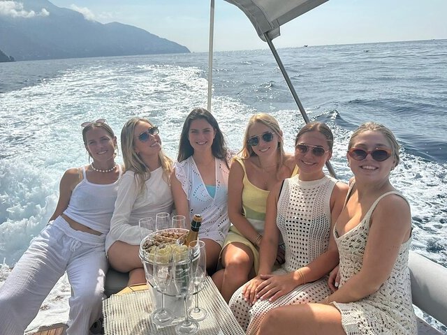 Six women sitting on a boat with ocean and mountains in the background, enjoying sunny weather.