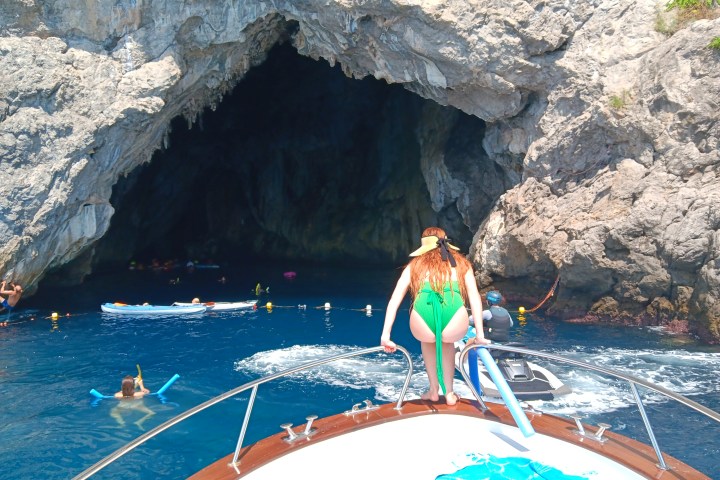 Person on boat near cave entrance with swimmers and kayaks in the blue water.