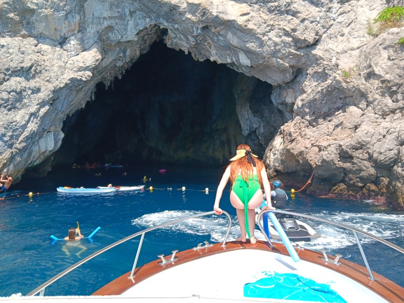 Person on boat near cave entrance with swimmers and kayaks in the blue water.