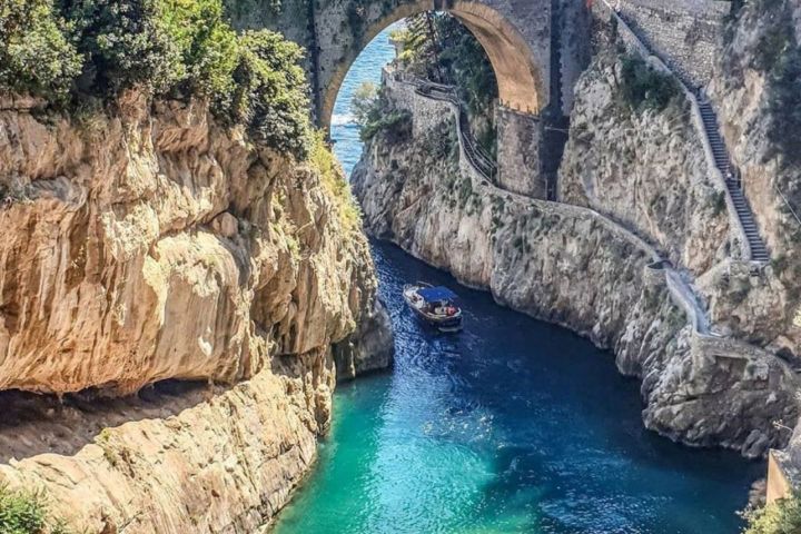 Stone bridge over a narrow cove with turquoise water and beachgoers.