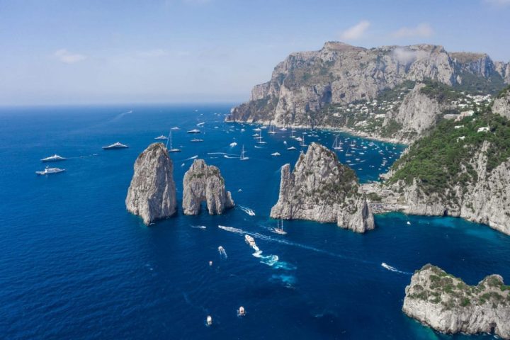 Aerial view of rocky island coast, sailboats, and yachts on blue ocean under clear sky.