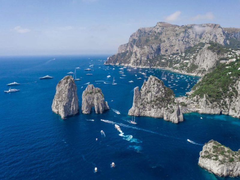 Aerial view of rocky island coast, sailboats, and yachts on blue ocean under clear sky.