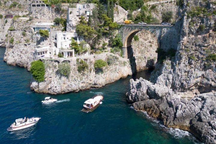 Cliffside village with arched bridge and boats in blue sea next to rocky coastline.