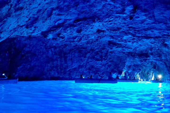 Boats in a cave illuminated by bright blue light reflecting on the water.