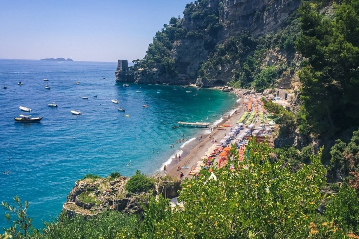 Scenic beach with colorful umbrellas, clear blue water, and lush cliffs in the background.