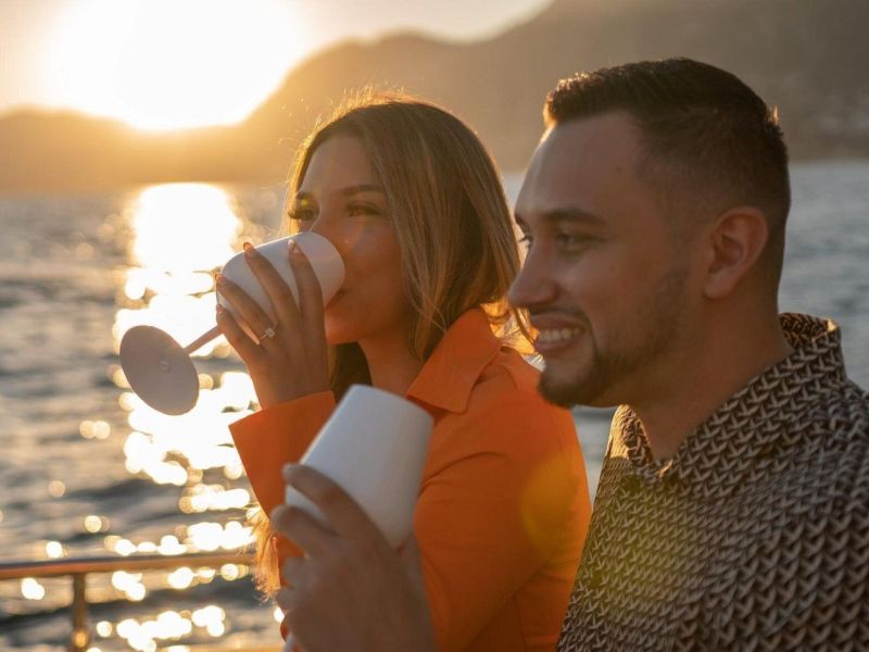 Couple on a boat at sunset, drinking from white cups, with the ocean and mountains in the background.