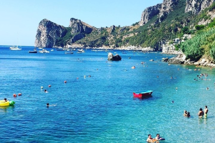 Clear blue sea with swimmers, boats, and rocky hills in the background on a sunny day.