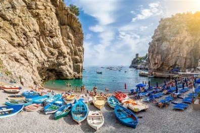 Seaside beach with colorful boats, sunbathers, cliffs, and umbrellas on a sunny day.