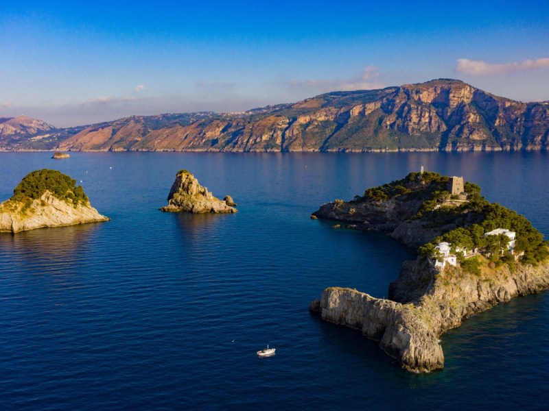 Aerial view of rocky islands with green foliage and a distant mountainous coastline under a clear blue sky.