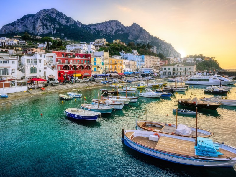 Boats in a vibrant harbor with colorful buildings and a mountain backdrop at sunset.