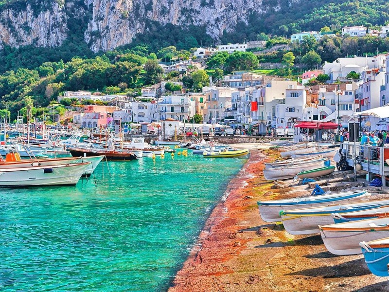 Harbor scene with colorful boats, turquoise water, and hillside town under a rocky cliff.