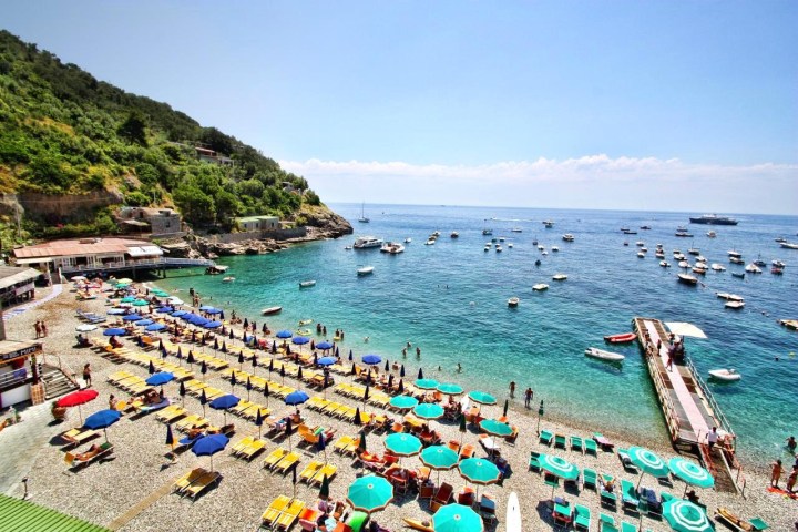 Crowded beach with colorful umbrellas and boats on a clear blue sea under a sunny sky.