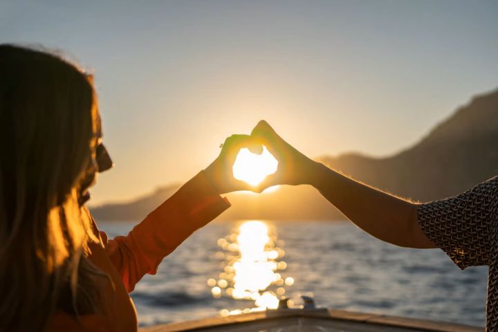 Two people forming a heart shape with hands against a sunset over water.