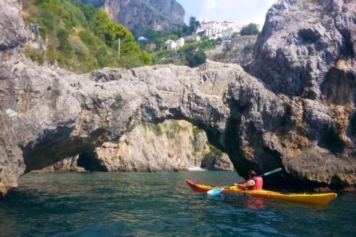 Person kayaking under a large rock arch on a sunny day.