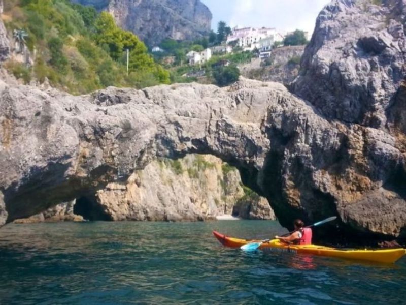 Person kayaking under a large rock arch on a sunny day.