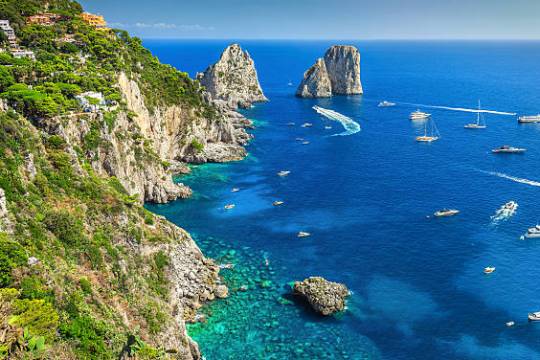 Scenic coastal view with cliffs, clear blue sea, boats, and distant rock formations on a sunny day.