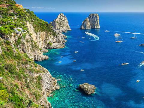 Scenic coastal view with cliffs, clear blue sea, boats, and distant rock formations on a sunny day.