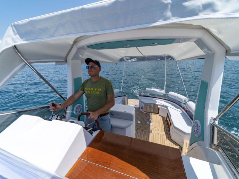 Man steering a boat with a canopy on a sunny day, surrounded by water.