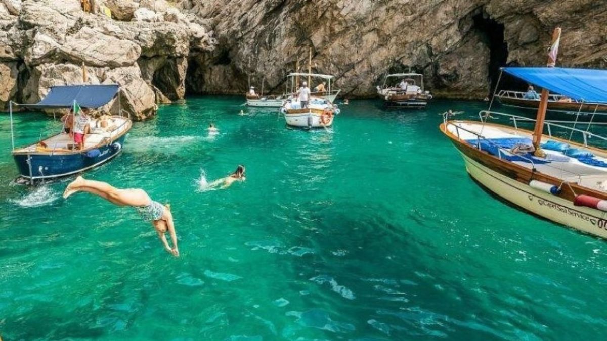 Person diving into turquoise water near boats and rocky cliffs.